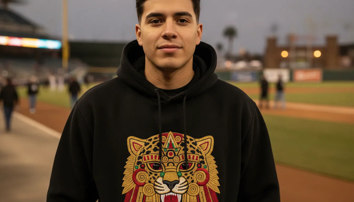 Hombre latino usando un hoodie negro premium con motivo de dios maya en un estadio de beisbol al atardecer